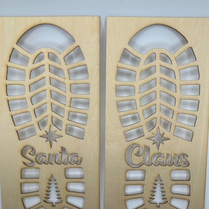 Può includere: Due decorazioni natalizie in legno con le parole "Santa Claus" ritagliate. Ogni pezzo presenta un ritaglio di un'impronta di stivale, una stella e un albero di Natale. Il legno ha una finitura chiara e naturale.
