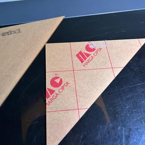 K&ouml;nnte beinhalten: Zwei klare, dreieckige Acrylteile. Eines tr&auml;gt den Text "EXILS". Das andere zeigt "MARGA CIPTA" in Rot, mit einem roten Logo. Die Acrylteile liegen auf einer schwarzen Oberfl&auml;che.