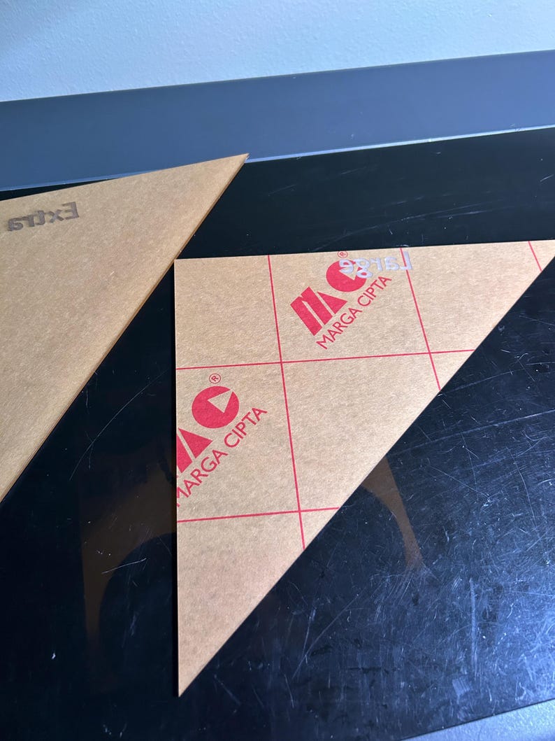 K&ouml;nnte beinhalten: Zwei klare, dreieckige Acrylteile. Eines tr&auml;gt den Text "EXILS". Das andere zeigt "MARGA CIPTA" in Rot, mit einem roten Logo. Die Acrylteile liegen auf einer schwarzen Oberfl&auml;che.