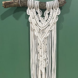 May include: A white macrame wall hanging with a natural wood branch at the top. The macrame is in the shape of a V with a series of circular patterns.