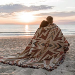 May include: A woven blanket with a geometric pattern in shades of brown, beige, and rust, fringed at the edges. The blanket is draped over two people on a sandy beach, with the ocean and a sunset in the background.