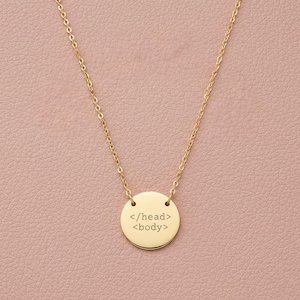 May include: A gold-colored necklace with a delicate chain and a round pendant. The pendant features the text "</head> <body>" in a simple, sans-serif font. The necklace is displayed against a soft pink background.