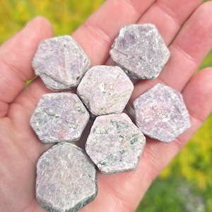 May include: A collection of seven rough-cut, hexagonal gemstones in varying shades of pink and gray, held in a hand. The stones have a natural, unpolished appearance, with visible textures and subtle green inclusions. The background is blurred, suggesting an outdoor setting.