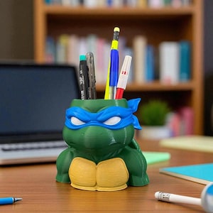 Può includere: Un portapenne verde e giallo a forma di personaggio delle Tartarughe Ninja, con una maschera blu. Il portapenne è pieno di penne e matite. Il portapenne è su una scrivania di legno.