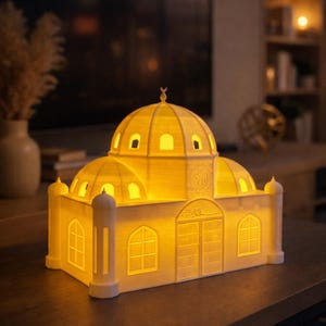 Puede incluir: Un modelo de mezquita blanco y brillante, con techo abovedado y minaretes. El edificio presenta ventanas y puertas arqueadas, con detalles intrincados y escritura árabe. El interior está iluminado, creando un ambiente cálido. El modelo está sobre una superficie de madera oscura.