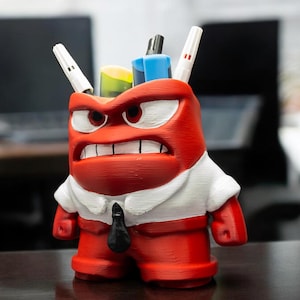 Könnte beinhalten: Roter und weißer 3D-gedruckter Schreibtisch-Organizer in Form einer wütenden Comicfigur mit schwarzer Krawatte. Der Organizer hat Stifte und Marker, die aus der Oberseite herausragen.