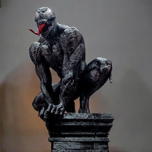 May include: A detailed sculpture of the Marvel Comics character Venom, in a crouching pose. The figure is primarily dark gray with red accents in the mouth and tongue. The sculpture is mounted on a dark gray, textured pedestal.
