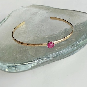 May include: A gold-toned cuff bracelet with a hammered texture and a vibrant, round, red gemstone. The open-ended design allows for easy wear. The bracelet is displayed on a clear, textured surface.