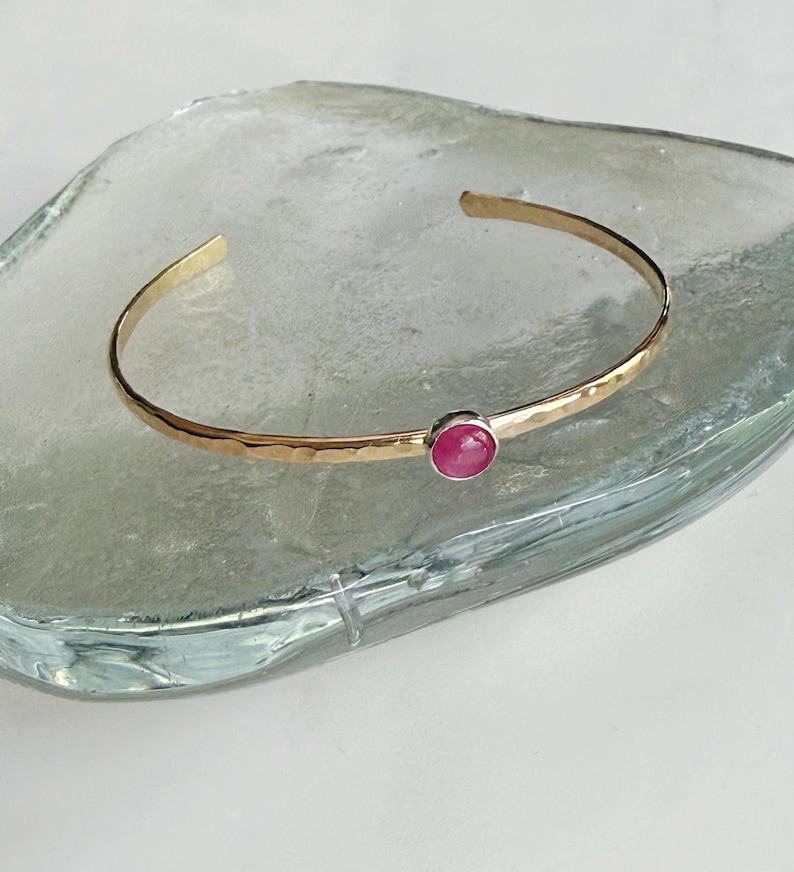 May include: A gold-coloured cuff bracelet with a hammered texture and a vibrant, round, red gemstone. The open design allows for easy wear. The bracelet is displayed on a clear, textured surface.