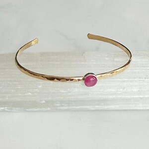 May include: Gold-coloured cuff bracelet with a hammered texture. It features a small, round, pink gemstone set in the centre. The open design allows for easy wear. A simple and elegant piece of jewellery.