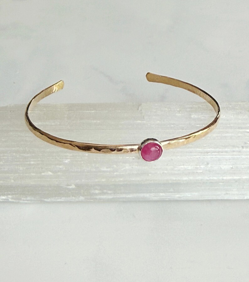 May include: Gold-coloured cuff bracelet with a hammered texture. It features a small, round, pink gemstone set in the centre. The open design allows for easy wear. A simple and elegant piece of jewellery.