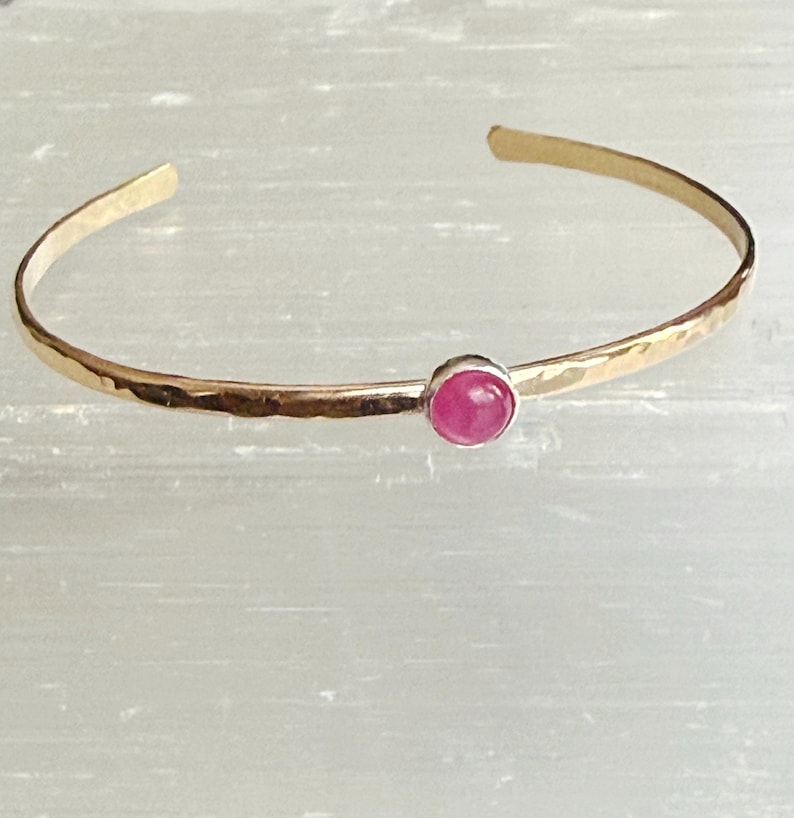 May include: Gold-coloured cuff bracelet with a hammered texture. The bracelet has a small, round, pink gemstone set in the centre. The open design allows for easy wear. A simple and elegant piece of jewellery.