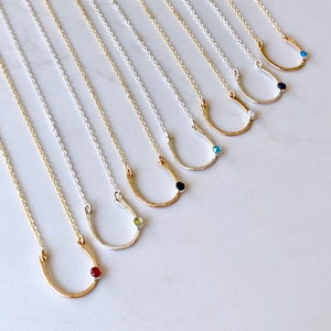 May include: A set of ten gold and silver horseshoe necklaces with different colored gemstones. The necklaces are arranged in a row on a white surface.