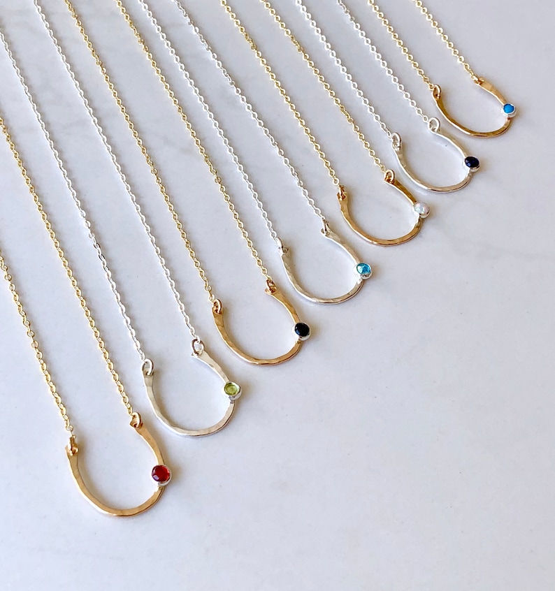 May include: A set of ten gold and silver horseshoe necklaces with different colored gemstones. The necklaces are arranged in a row on a white surface.