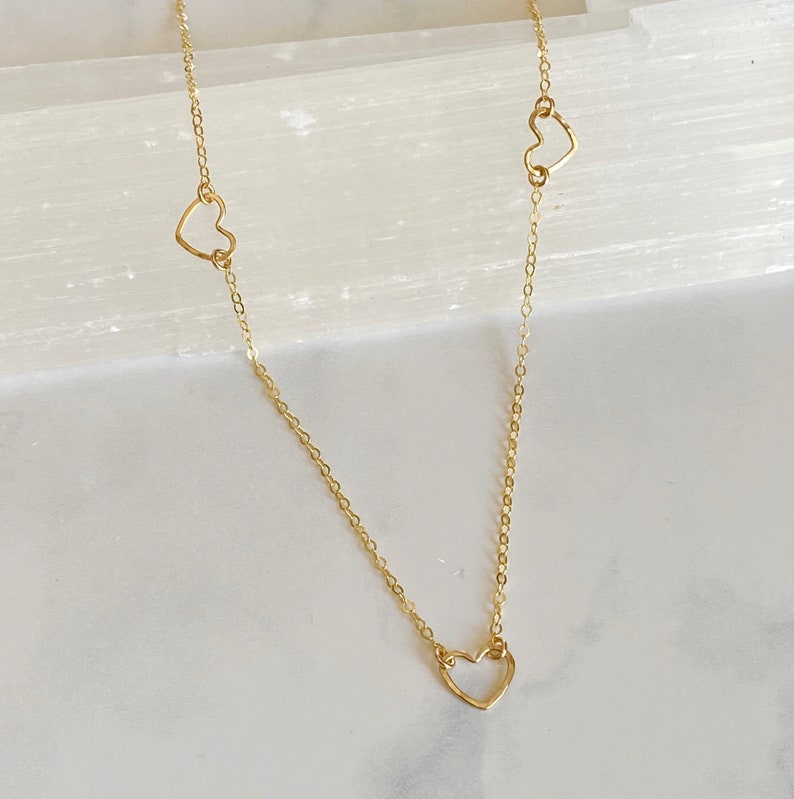 May include: A gold chain necklace with three delicate open heart charms. The necklace is on a white background.