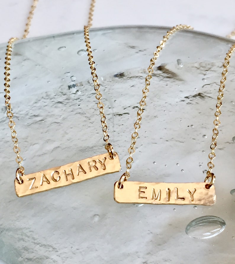 May include: Two gold necklaces with personalized nameplates. The top necklace has the name "ZACHARY" on the nameplate. The bottom necklace has the name "EMILY" on the nameplate.