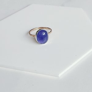 May include: A ring featuring a smooth, oval-shaped purple gemstone set in a silver bezel. The band of the ring is made of thin, textured gold metal with a hammered finish, creating a contrast between the polished stone and the metallic band.