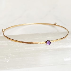 May include: A delicate gold bangle bracelet with two small amethyst gemstones. The bracelet is hammered for a textured look.