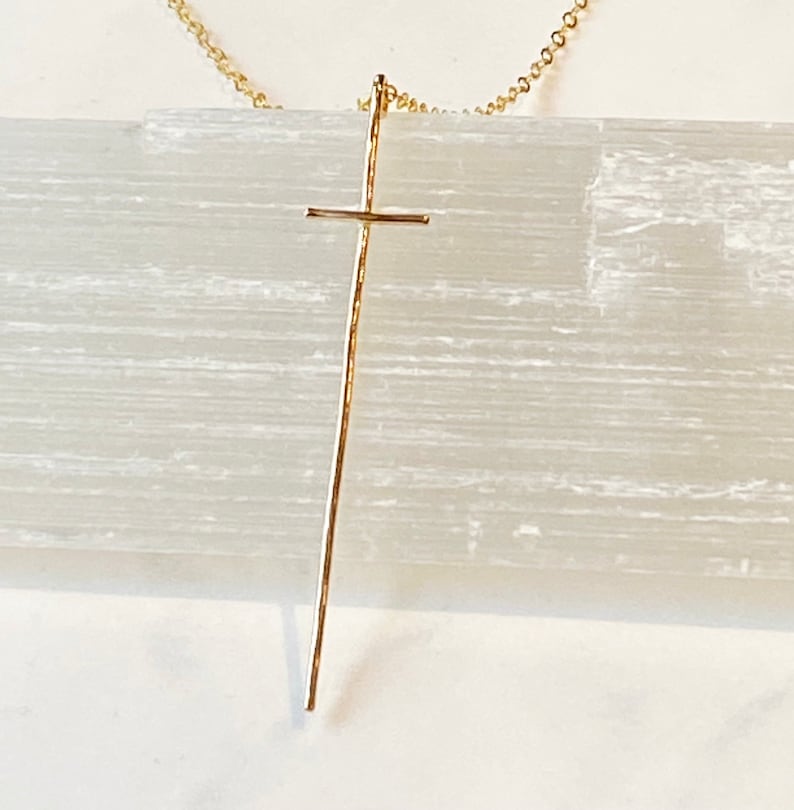 Pode incluir: Um colar dourado com um pingente de cruz longo e fino.