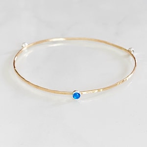 May include: A gold-coloured bangle bracelet with a hammered texture. The bracelet features a small, round, bright blue gemstone set in a silver-coloured bezel. Two small silver-coloured beads are also on the bracelet.