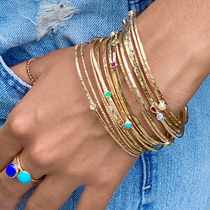 May include: A stack of gold bangles with small gemstones set in each band. The bangles are worn on a person's wrist. The gemstones are various colors, including blue, green, red, and white.