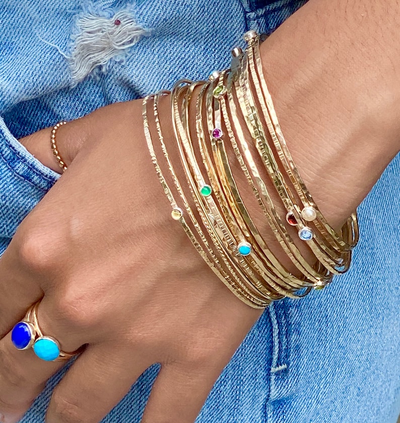May include: A stack of gold bangles with small gemstones set in each band. The bangles are worn on a person's wrist. The gemstones are various colors, including blue, green, red, and white.