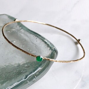 May include: A delicate gold bangle bracelet with a textured finish and two small green gemstones.