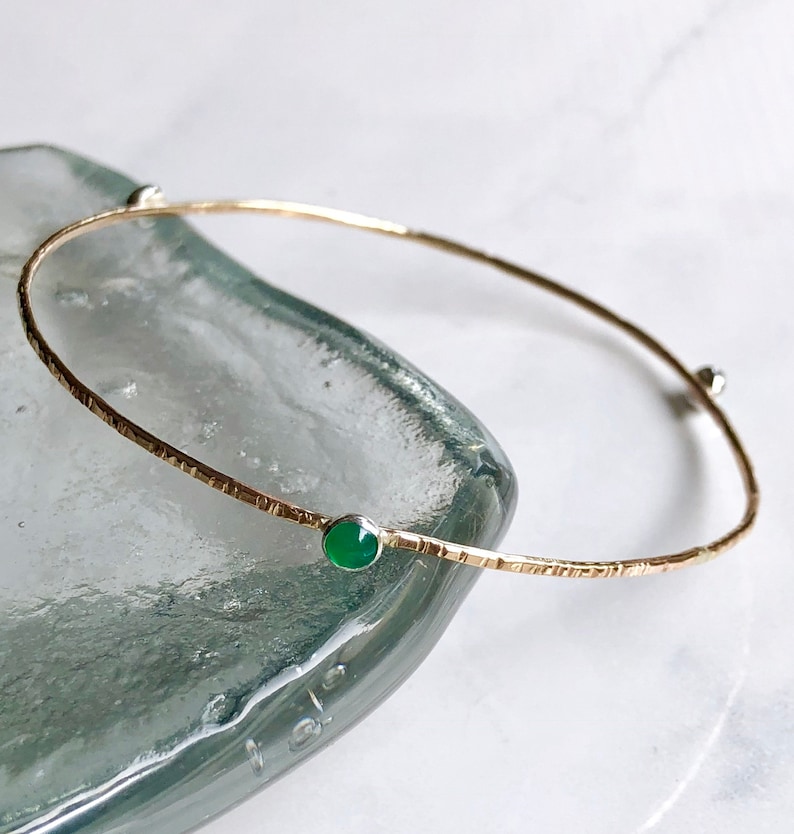May include: A delicate gold bangle bracelet with a textured finish and two small green gemstones.