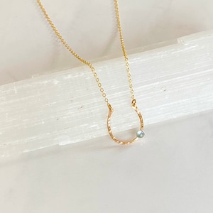 May include: A gold necklace with a hammered horseshoe pendant and a light blue gemstone.