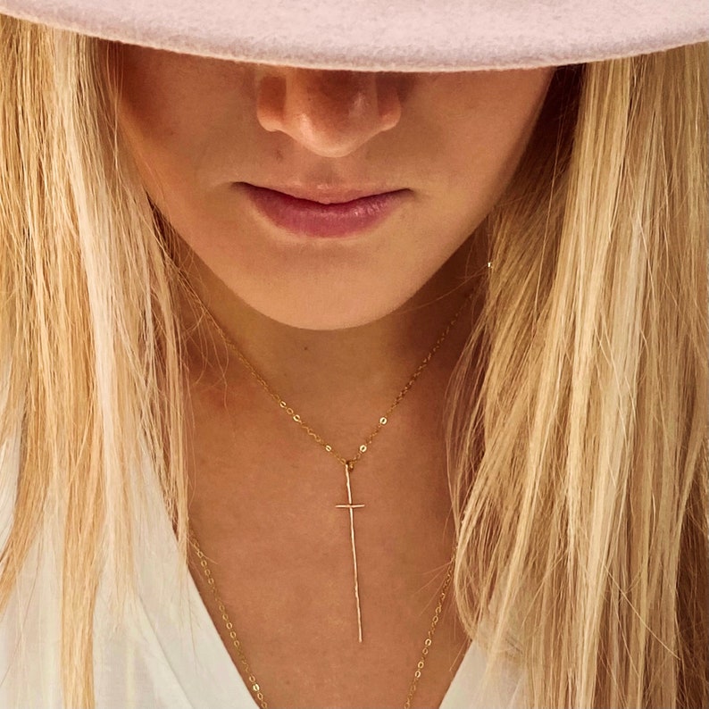 Pode incluir: Um close-up de uma mulher usando um colar de ouro com um pingente de cruz longo e fino. O colar est&aacute; sobreposto a uma segunda corrente mais curta. A mulher est&aacute; usando uma camisa branca e tem cabelo loiro.