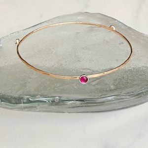 May include: A gold-coloured bangle bracelet with a hammered finish. It has a small, round, bright pink gemstone set in a silver bezel. The bracelet is displayed on a clear, textured surface.