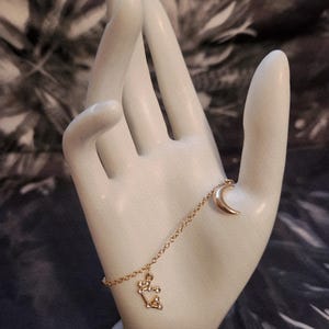 May include: A gold-colored bracelet featuring a crescent moon charm and a constellation charm. The bracelet is displayed on a white hand-shaped sculpture against a blurred background.