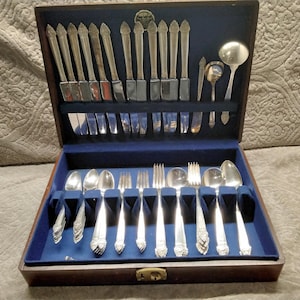 May include: A wooden flatware chest with a blue velvet interior containing a set of silver-toned utensils. The collection includes knives, forks, and spoons with decorative handles, organized in two tiers within the hinged storage box.