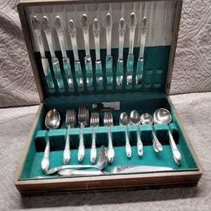 May include: A silver-toned flatware set housed in a wooden storage case with a green velvet interior. The set includes various knives, forks, and spoons with decorative handles, arranged in dedicated slots within the box for organized storage.