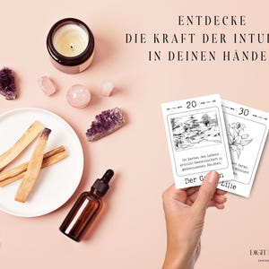 Hand hält Lenormand-Karten "Der Garten" und "Die Lilie," präsentiert mit spirituellen Accessoires wie Palo Santo und Kristallen.