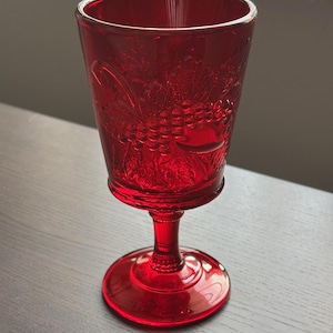May include: A ruby red glass goblet with a detailed grape and leaf pattern. The stemware features a round base and a slender stem, perfect for serving wine or other beverages. The glass is approximately 6 inches tall.