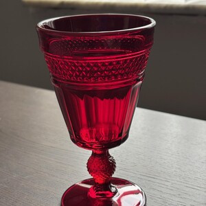 May include: A ruby red glass goblet with a textured design. The goblet features a round base, a decorative stem, and a patterned rim. The glass is transparent, allowing light to pass through.