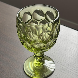May include: A green-tinted glass goblet with a textured, patterned bowl and a round base. The stem is short and octagonal. The goblet is sitting on a dark wooden surface.