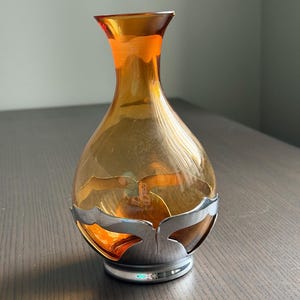 May include: A translucent amber glass vase with a flared neck and a rounded body, held within a polished silver-toned metal base featuring a decorative cutout design that wraps around the lower portion of the glass vessel.
