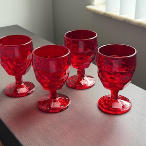 May include: A set of four ruby red glass goblets featuring a faceted, diamond-like geometric pattern on the bowl. Each goblet stands on a short, sturdy stem with a circular base. Small white rectangular labels are attached to the base of each glass.