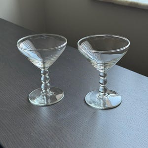May include: Two clear, stemmed cocktail glasses with a classic coupe shape. The stems feature a series of small, round glass beads. The glasses sit on a dark gray surface, with a window in the background.