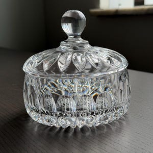 May include: A clear glass candy dish with a matching lid featuring a spherical knob handle. The bowl and lid are decorated with a repeating vertical arched pattern and a scalloped rim, creating a textured, reflective surface throughout the piece.
