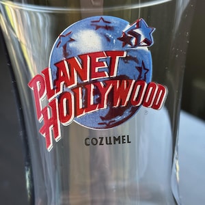 May include: Clear glass Planet Hollywood souvenir glass with a flared shape. The logo features a blue background with red text and a star design. The word "COZUMEL" is printed below the logo.