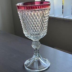 May include: A clear glass goblet with a diamond-cut pattern on the bowl and a red rim. The goblet has a decorative stem and a wide, circular base. The glass is transparent, allowing light to pass through.