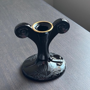 May include: Black ceramic candle holder with gold accents and a floral design. The holder has two curled arms and a wide base.