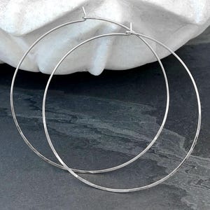 May include: A pair of large, thin, circular silver hoop earrings. Each earring features a simple, continuous wire design with a small, integrated post closure at the top, creating a clean and minimalist aesthetic for these metallic jewelry pieces.