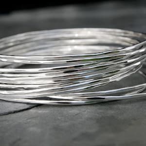 May include: A stack of multiple thin, circular silver bangles. Each bangle features a reflective, polished surface with a subtle hammered texture that creates small, irregular indentations along the circumference of the metal bands.
