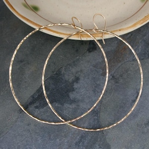 May include: A pair of large, circular hoop earrings crafted from thin, hammered gold-tone metal. Each earring features a delicate wire hook closure at the top, with a textured, reflective surface that catches the light along the entire circumference.