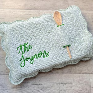 May include: A light green quilted placemat with a scalloped edge. The placemat has a diamond pattern and features the embroidered text "The Joyners" in green script. A wooden spoon and a small wooden T-shaped object are attached.
