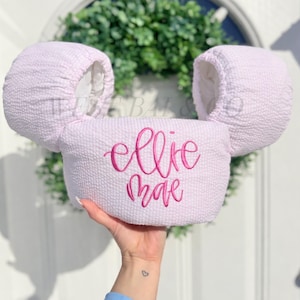 May include: A light pink, textured fabric mouse ear headband with the name "ellie mae" embroidered in a bright pink script. The headband has two large, rounded ears and a central band.